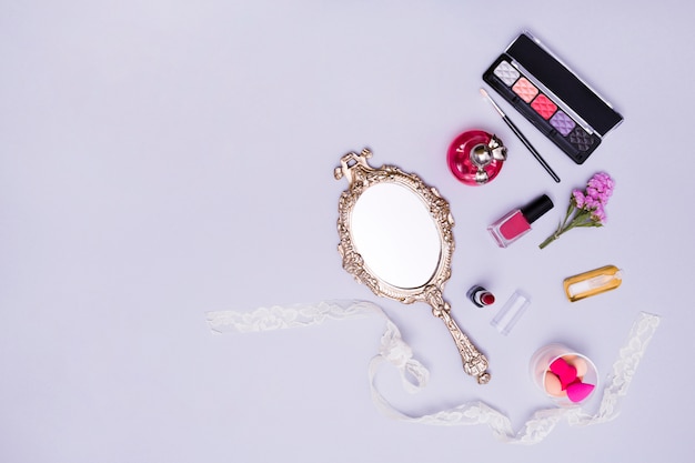 Vintage hand mirror; lipstick; nail varnish; sponge; perfume bottle and eyeshadow palette on purple background