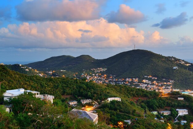 버진 아일랜드 세인트 토마스(Virgin Islands St Thomas)는 다채로운 구름, 건물 및 해변 해안선이 있는 일몰 산의 전망을 제공합니다.