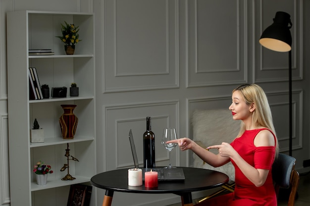 Virtual love cute blonde girl in red dress on distance date with wine and candle pointing at camera