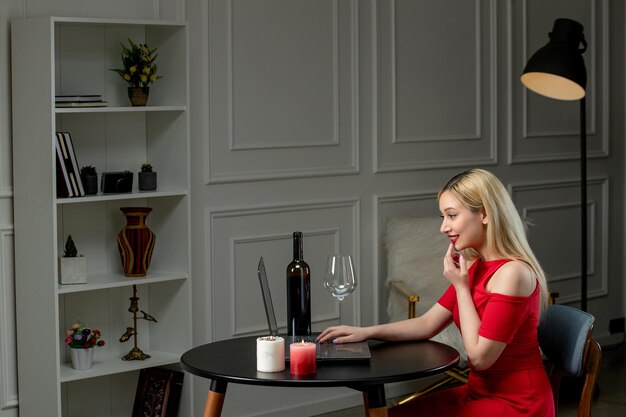 Virtual love cute blonde girl in red dress on distance date with wine and candles finger on face