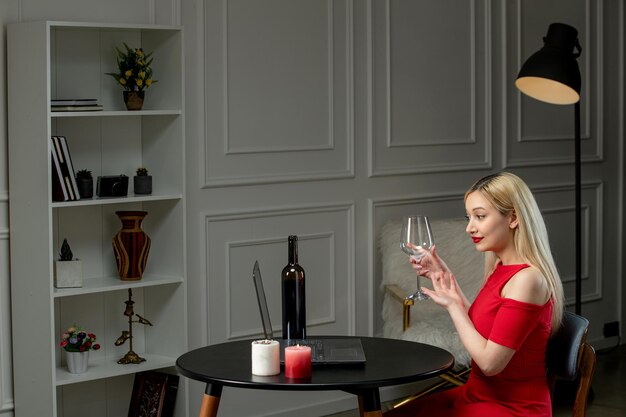 Virtual love cute blonde girl in red dress on distance date with wine and candles holding glass