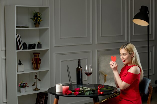 Virtual love cute blonde girl in red dress on distance date with wine and candles holding heart