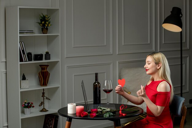 Virtual love cute blonde girl in red dress on distance date with wine and candles with heart sign