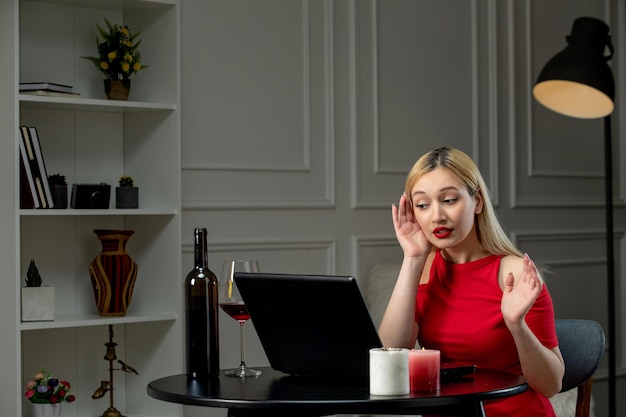 Virtual love cute blonde girl in red dress on distance date with wine concentrating to listen