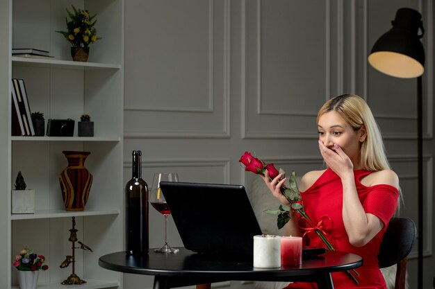 Virtual love cute blonde girl in red dress on distance date with wine covering face with flowers