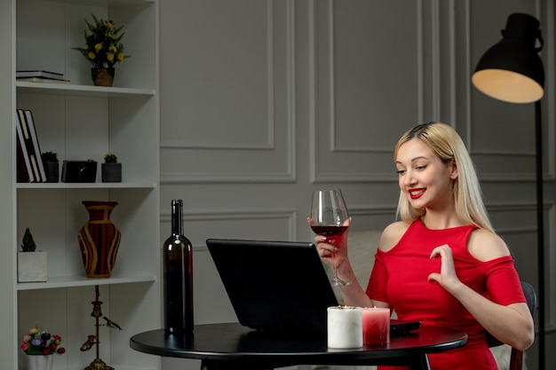 Virtual love cute blonde girl in red dress on distance date with wine holding heart sign