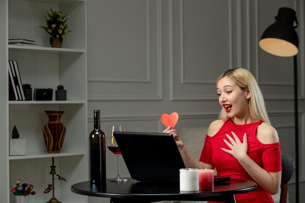 Virtual love cute blonde girl in red dress on distance date with wine holding red heart paper