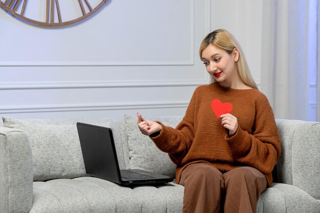 가상 사랑의 귀여운 젊은 금발 소녀는 손가락 심장을 잡고 있는 원거리 컴퓨터 날짜에 아늑한 스웨터를 입고 있습니다.