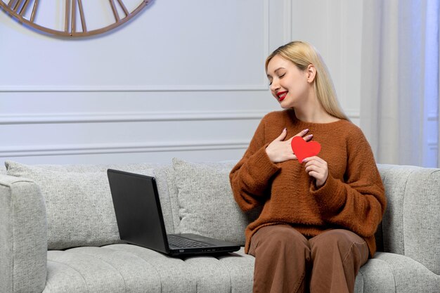 Virtual love cute young blonde girl in cozy sweater on distance computer date holding paper heart