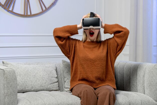 Virtual love cute young blonde girl in cozy sweater wearing virtual d goggles holding head shocked