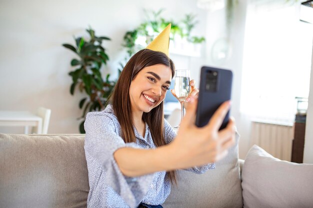 Virtual Party Happy young woman in hat having video conference online meeting with friends and family holding glass of wine toasting and celebrating birthday staying at home