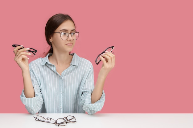 Vision, optics, eyesight, myopia, people and eyewear concept. Picture of stylish beautiful short sighted young female choosing distance glasses, holding two pairs of spectacles, having indecisive look