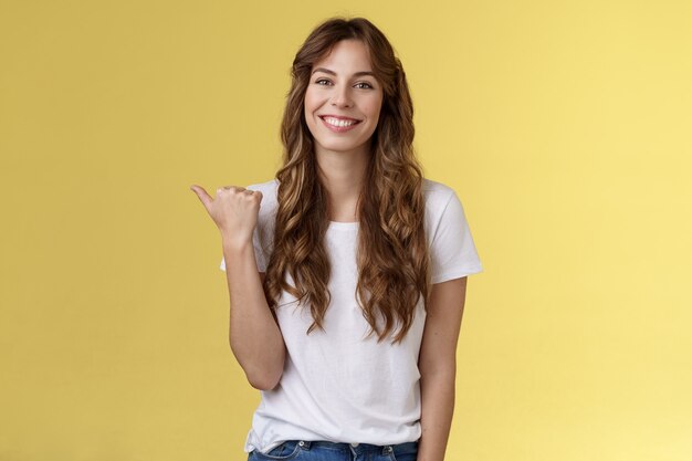 Visit see yourself. Cheerful charismatic good-looking outgoing girl long curly haircut showing place do good hairstyle smiling happily delighted pointing thumb left introduce promo yellow background