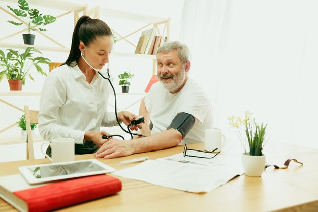 The visiting nurse or health visitor taking care of senior man. Lifestyle portrait at home. Medicine, healthcare and prevention. Girl checking or measuring patient's blood pressure during the visit.