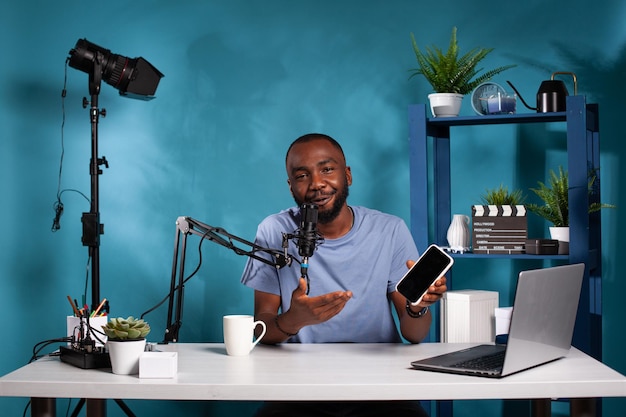 Vlogger announcing new giveaway on social media presenting smartphone from sponsor while talking into microphone. Famous influencer recording online contest in vlogging studio.