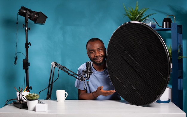 Vlogger explaining features of studio flash light modifier sitting at desk with microphone in vlogging studio. Portrait of photography equipment reviewer presenting beauty dish honeycomb grid.