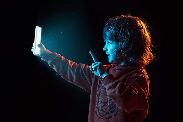 Vlogging with smartphone. Caucasian boy's portrait on dark studio background in neon light. Beautiful curly-haired model. Concept of human emotions, facial expression, sales, ad, modern tech, gadgets.