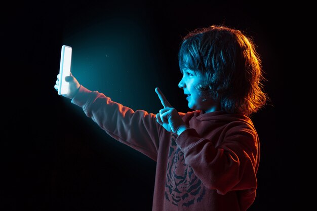 Видеоблог со смартфона. Портрет кавказского мальчика на темном фоне студии в неоновом свете. Красивая кудрявая модель. Концепция человеческих эмоций, выражения лица, продаж, рекламы, современных технологий, гаджетов.