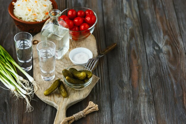 Vodka with salted vegetables on wooden surface. Alcohol pure craft drink and traditional snack, tomatoes, cabbage, cucumbers. Negative space. Celebrating food and delicious.