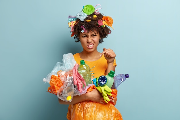 Voluntary free assistance. Irritated black woman worried with environmental pollution problem, carries trash bag with recycled plastics, clenches teeth from annoyance, isolated over blue wall