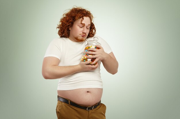 Voracious greedy fat man with red curly hair holding jar of sweets tight