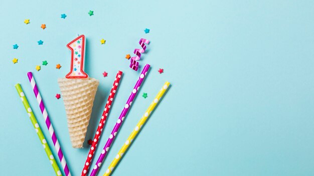 Waffle cone with number one candle; streamer and drinking straws on blue background