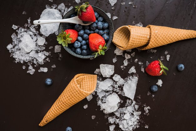 Waffle cones near fresh berries on dish and spoon among ice