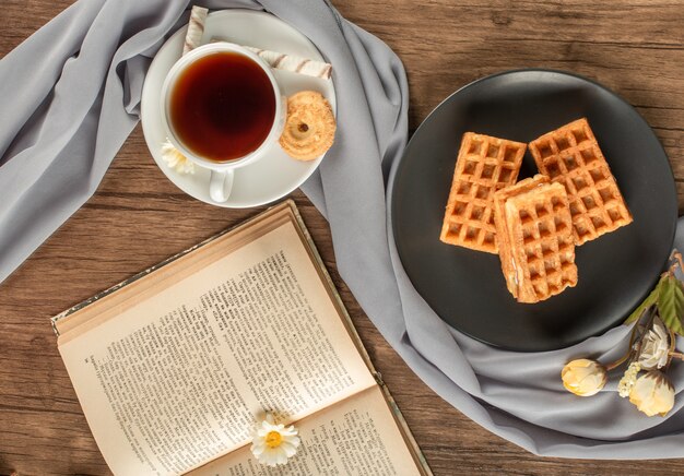 Waffles in a black saucer, a cup of tea and a book. top view