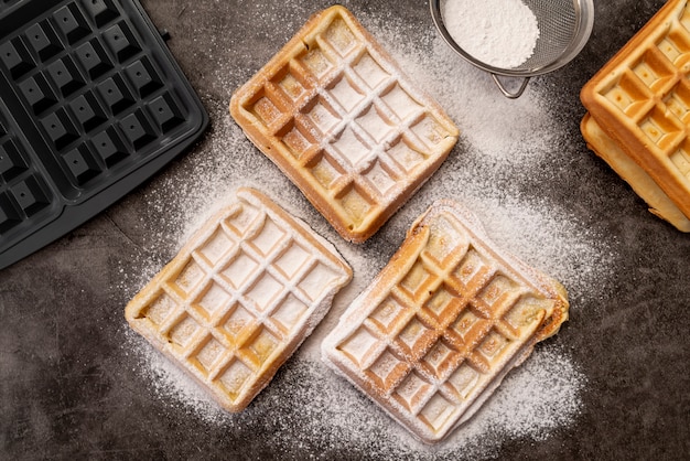 Waffles covered in powdered sugar with waffle maker and sieve