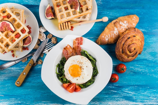 Waffles with fig; baked pastries and egg fried egg on white plates over blue textured backdrop