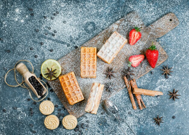 Waffles with spices, cookies, choco chips, strawberry, strainer flat lay on grungy and cutting board surface