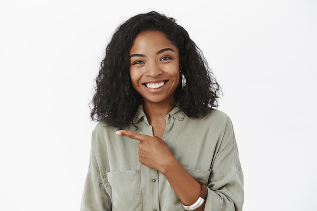 Foto gratuita mezzo busto di donna affascinante dalla pelle scura dall'aspetto amichevole divertito con l'acconciatura riccia in camicia grigia che punta a sinistra sorridendo gioiosamente