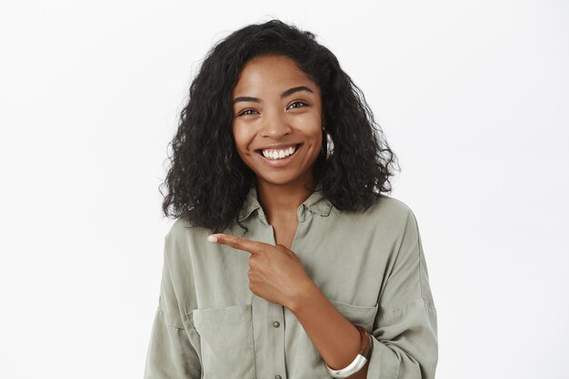 Waist-up of amused friendly-looking charming dark-skinned woman with curly hairstyle in grey shirt pointing left smiling joyfully