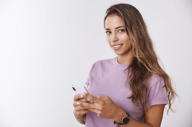 Splendida ragazza sorridente felice in vita che tiene in mano uno smartphone che sembra felice e tenera macchina fotografica, pensando di catturare sotto selfie pubblicando foto sui social network, chiedendoti di scattare foto sul muro bianco