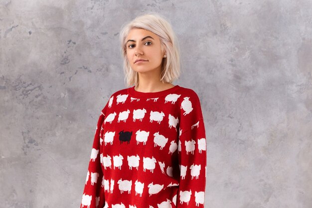 Waist up image of trendy young female with dyed messy bob hairstyle posing in red jumper with white lambs, isolated at blank  wall with copyspace for your advertising text or information