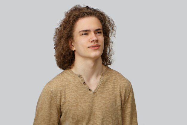 Waist up isolated picture of handsome young male with loose curly hairdo and smooth clean face looking sideways with thoughtful dreamy smile. Human facial expressions, emotions and feelings