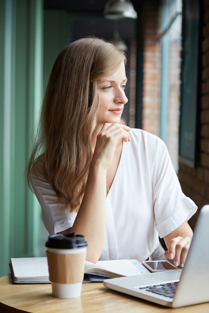 Waist up of pensive young woman sitting at desk looking in the window