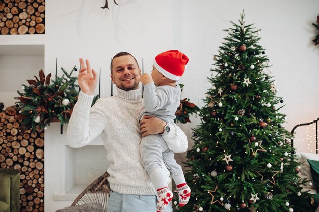 Waist-up photo of a happy dad waving and holding a child near Christmas tree