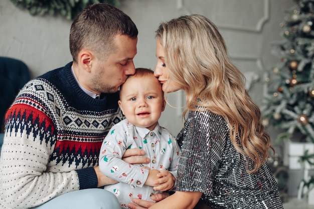 愛する両親がクリスマスツリーで赤ちゃんの頭にキスをしているウエストアップ写真