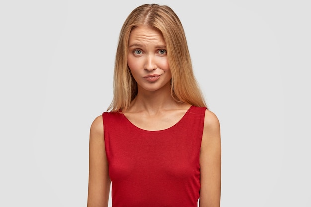 Waist up portrait of adorable female with hesitant facial expression, has doubtful look, feels concerned, wears red vest, has healthy skin, isolated over white wall. People and beauty concept