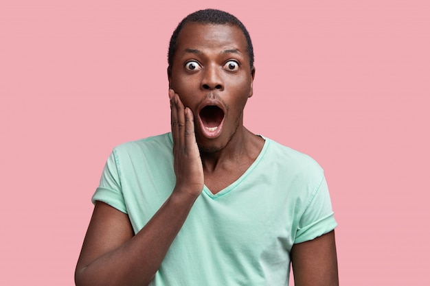 Free photo waist up portrait of amazed bugged eyed dark skinned male in casual t shirt, being surprised to hear shocking news, isolated over pink