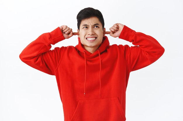 Waist-up portrait of annoyed young man, asian male student complaining on loud neighbours, shut ears and grimacing, looking up irritated, hear loud annoying music, white wall.