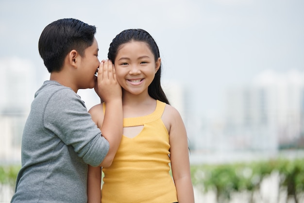 Waist up portrait of Asian kids looking at camera, boy whispering a secret to his gorlfriend