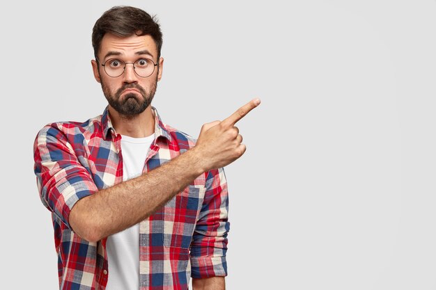 Waist up portrait of astonished Caucasian male purses lips and looks surprisingly, points at upper right corner, wears fashionable outfit, stands against white wall with copy space for text