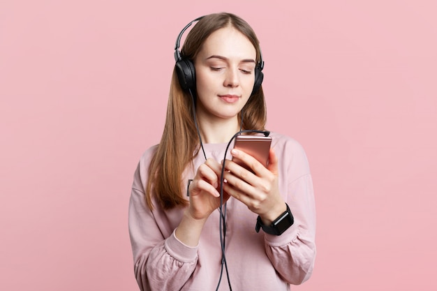 Waist up portrait of beautiful brunette female wears headphones, checks song from playlist