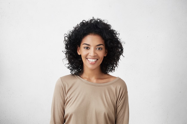 Free photo waist up portrait of casually dressed young mixed race female with curly hair smiling cheerfully during audition for role in tv series, feeling excited and a big nervous, trying to impress director
