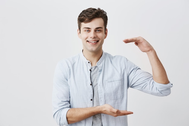 Waist-up portrait of caucasian male model joyfully smiling with white teeth, demonstrating size of the box with present for his girlfriend, gesturing actively. Body language