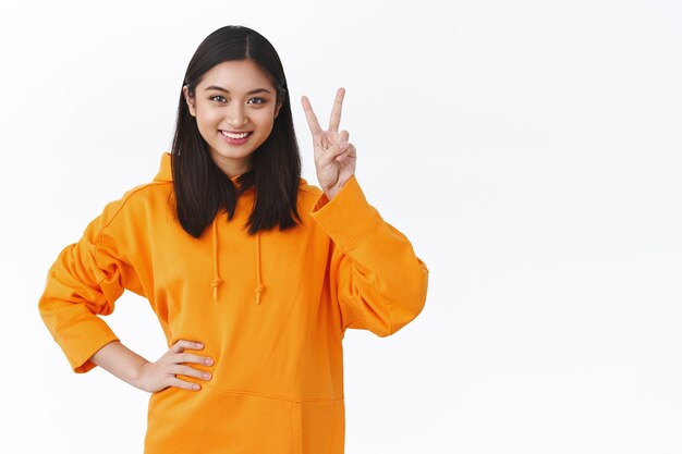 Waist-up portrait cheerful young asian girl showing kawaii peace sign and smiling optimistic, standing confident and ready for action, tackle any task, moving up career stairs, white wall