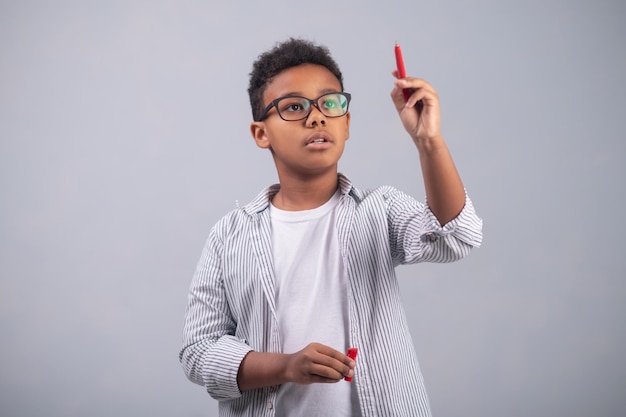 Waist-up portrait of a concentrated bespectacled schoolkid holding a pen in his raised hand and thinking