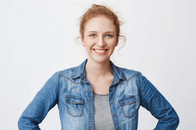 Waist-up portrait of cute happy european redhead model with hair combed in bun holding hands on waist